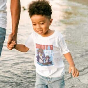 A child wearing a white color wubba t-shirt with text that says, Daddys First Mate with a cute cartoon illustration of and papa and son penguin sailors, with the little penguin saluting the papa who is also the captain of the crew